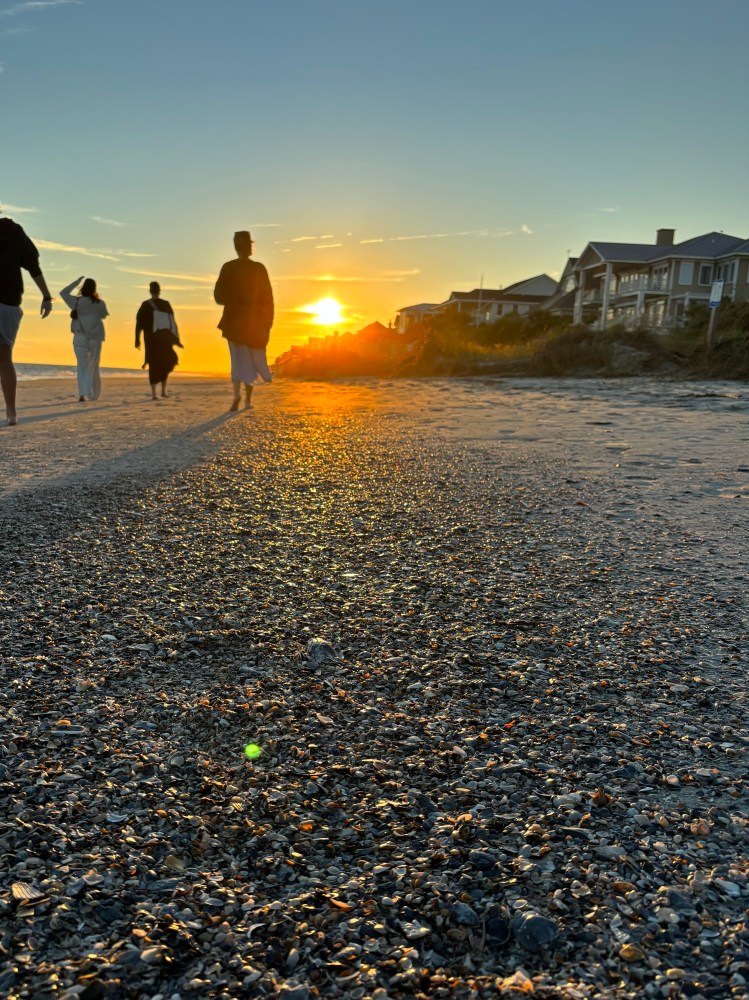 People walking on a beach at sunset with houses in the background.